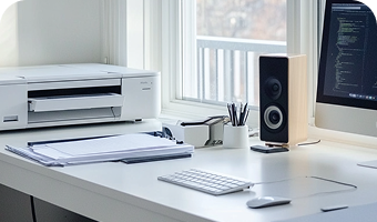 Ein heller Arbeitsplatz mit Computer, Drucker und einem Lautsprecher, direkt vor dem Fenster.